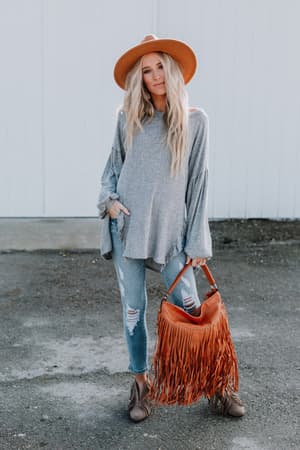 A woman with long, wavy blonde hair and a slight smile stands outdoors wearing the Charlie Cozy Top - Gray, ripped jeans, a wide-brim hat, brown shoes, and holding a brown fringed handbag.