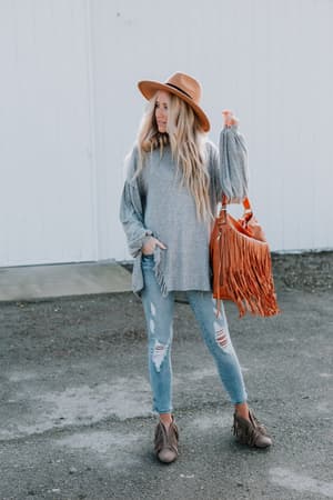A woman with long, wavy blonde hair poses against a white background in the Charlie Cozy Top - Gray, brown hat, puff long sleeves, ripped jeans, brown ankle boots, and holds an orange fringe bag.