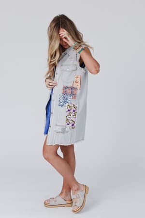 A woman models the One Of A Kind Sleeveless Denim Dress Vest - Denim Multi, adorned with colorful patches. She pairs it with beige sandals and layered bracelets, tilting her head down to partially cover her face with her hand against a plain background.