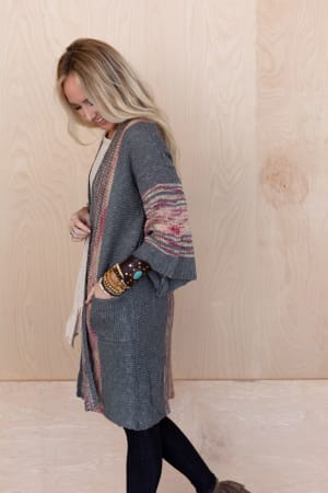 A woman with long blonde hair wears the Rose Boho Throw On Cardigan in gray, styled over a light top and dark pants with brown shoes, standing sideways against a light wooden wall and looking down with one hand near her face.