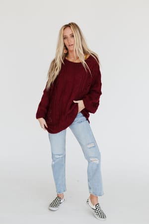 A woman with long blonde hair wears the Golden Hour Bubble Sleeve Sweater Dress in burgundy, standing against a plain white background and posing relaxedly for the camera.