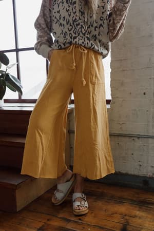 A person wearing the Relaxing Robin Wide Leg Pant in mustard stands indoors near a brick wall and large window, with one foot on a step.