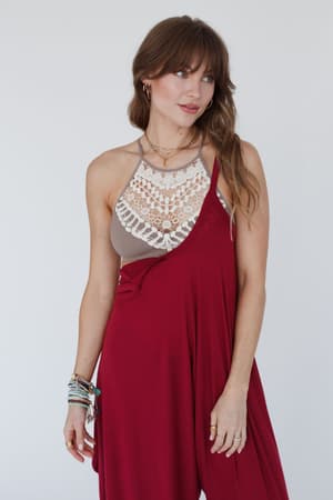A woman with long brown hair wears a red asymmetrical dress over the Crochet High Neck Bralette - Mocha, accessorized with layered necklaces and bracelets, standing against a plain white background.