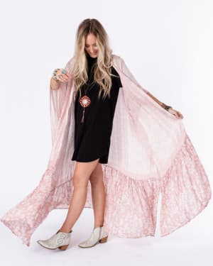 A woman with long blond hair wears a black dress, white ankle boots, and the Paisley Tapestry Free Flow Duster Kimono in Blush as she poses against a white background, looking down and smiling.