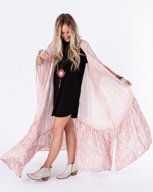 A woman with long blond hair wears a black dress, white ankle boots, and the Paisley Tapestry Free Flow Duster Kimono in Blush as she poses against a white background, looking down and smiling.
