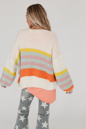 A person with long, blonde hair faces away, wearing the Retro Babe Striped Cardigan - Cream—an oversized knit with pastel stripes—over a peach shirt and gray flared pants with white stars.