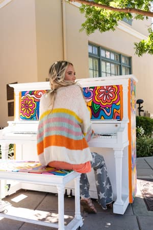A woman with long blonde hair wearing the Retro Babe Striped Cardigan - Cream and star-patterned pants plays a white piano decorated with vibrant floral designs outdoors.