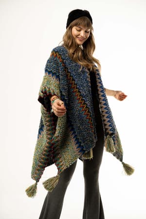 A woman wears the Desert Dreams Crochet Ruana – Cobalt, showcasing colorful zigzag patterns and large tassels, paired with wide-leg gray pants and a black headband—a perfect boho chic look.