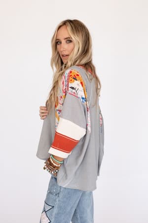 A woman with long blonde hair models the Britt Patchwork Top in heather gray, featuring 3/4 length colorful patterned sleeves and an oversized fit. She pairs it with blue jeans and layered beaded bracelets, standing sideways and looking at the camera.