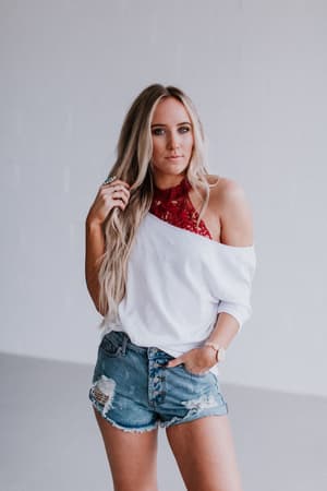 A woman with long blonde hair wears a white off-shoulder top, distressed denim shorts, and the Baroque High Neck Velvet Bralette in Wine as she stands against a plain light background, gazing at the camera with a relaxed expression.