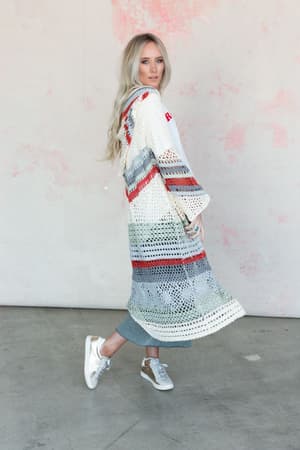 A woman with long blonde hair wears the Love In Layers Cardigan - Multi, a bohemian duster-length crochet piece, over a white top and blue jeans. She poses on a concrete floor against a light pink and white textured wall.