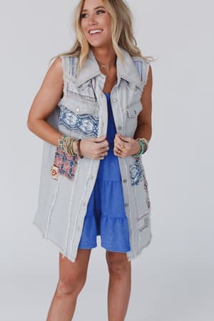 A woman with long blonde hair and stacked bracelets smiles in the One Of A Kind Sleeveless Denim Dress Vest - Denim Multi, featuring patchwork denim and colorful embroidered patches, standing against a plain light background.