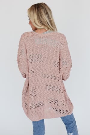 A woman with blonde hair, seen from the back, wears the Cara Oversized Cardigan in pink over a gray top and blue jeans—a perfect transitional layering piece—against a plain white background.