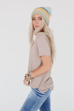 A woman with long blonde hair wears a colorful striped beanie, blue jeans, multiple beaded bracelets, and the Scoop Neck Bralette Tee in Khaki, standing sideways against a plain white background.