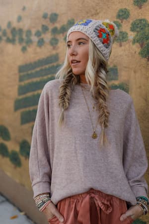 A woman with long blonde hair in fishtail braids wears The Nest Fly With Me Batwing Sleeve Knit Sweater in Sand, a floral knit hat, and loose pants as she stands before a green mural, gazing to the side.
