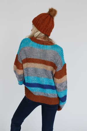 A person wears the Go For It Striped Sweater - Blue with blue, brown, gray, and beige stripes, paired with dark jeans and a rust beanie topped with a fur pom-pom, standing against a plain white background facing away.