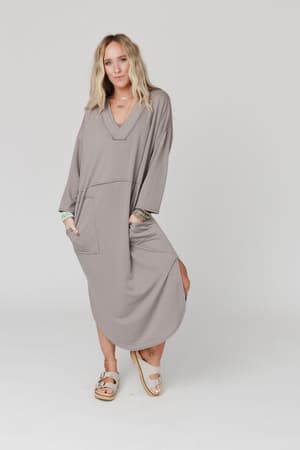 A woman with long blonde hair wears The Nest Meadow Long Sleeve Slit Maxi Dress - Coco, a relaxed-fit gray maxi dress with front pockets and side slit. She stands barefoot in white sandals against a plain white background.