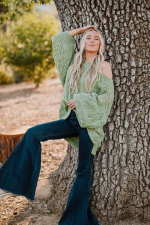 A woman with long blond hair in the Golden Hour Bubble Sleeve Sweater Dress in light green leans against a tree outdoors, smiling with closed eyes as sunlight filters through the greenery behind her.