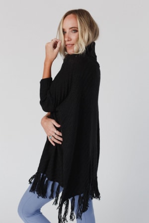 A woman with shoulder-length blonde hair wears the Paloma Poncho Sweater - Black, featuring an oversized silhouette and fringe detail, over light blue pants. She stands sideways, looking at the camera against a plain white background.