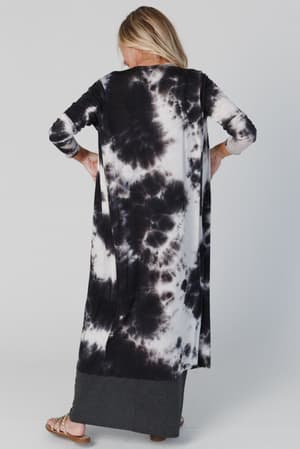 A woman faces away, wearing the My Go To Duster Cardigan in black tie dye over a dark gray skirt with sandals. Her head is turned slightly left, showing her blonde hair. The background is light and plain.