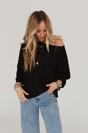 A woman with long blonde hair wears the Fly With Me Batwing Sleeve Knit Sweater in black, paired with light blue jeans and multiple bracelets, looking down with one hand in her pocket against a plain white background.
