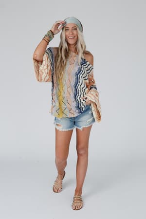 A smiling woman with long blonde hair models the Rainbow Shores Knit Sweater - Multi, featuring a colorful boho design, slouchy loose fit, and flare sleeves, paired with ripped denim shorts and accessories against a plain white background.
