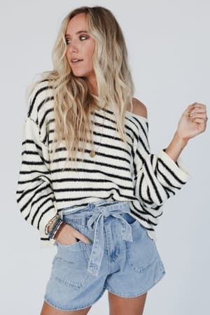 A woman with long, wavy blonde hair wears the All My Stripes Oversized Sweater in cream and black off one shoulder, paired with high-waisted, tie-front denim shorts, standing against a plain light background.