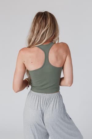 A woman with long, wavy blonde hair is seen from behind, wearing the olive High Neck Racerback Brami and loose light gray pants against a plain light background.