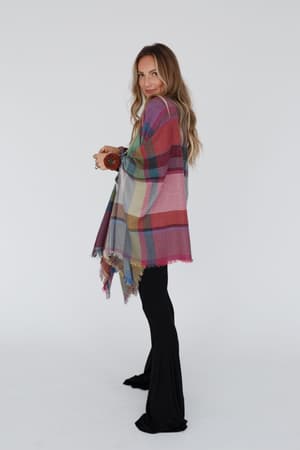 A woman with long hair stands sideways, smiling, wearing the Cozy Plaid Pocket Ruana Wrap - Fuchsia over a black top and flared black pants, set against a plain white background.