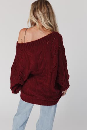 A woman with long blonde hair wears the Golden Hour Bubble Sleeve Sweater Dress in burgundy, styled off one shoulder, standing with her back to the camera against a plain backdrop.