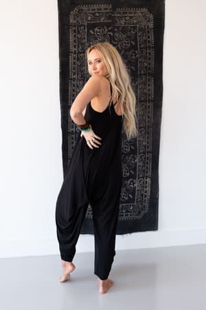 A woman with long blonde hair poses barefoot in The Perfect Harem Jumpsuit - Black, standing before a decorative black and white tapestry on a white wall.