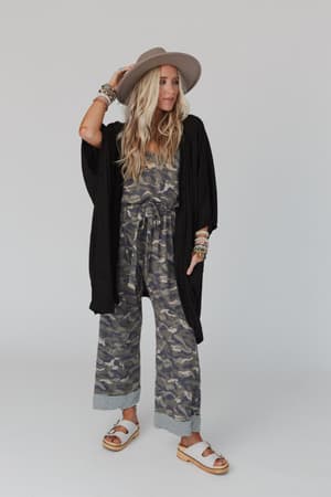 A woman stands confidently against a light backdrop, layering bracelets and necklaces over her look—a black kimono, camouflage jumpsuit, platform sandals, and The Nest Boho Essential Hooded Ribbed Sweater draped stylishly off one shoulder.