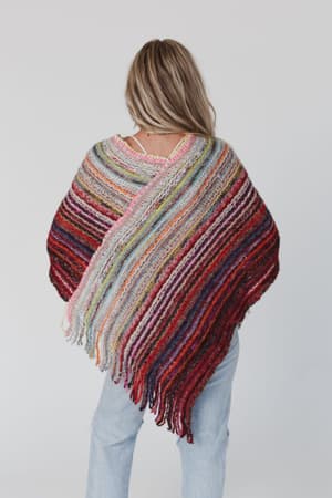 A person with long, light brown hair is shown from the back wearing the Madeleine Poncho - Multi, a colorful knit with fringe edges, paired with light blue jeans against a plain, light background for a boho look.