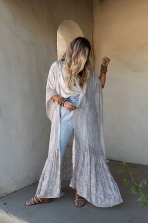 A woman with long, wavy blonde hair stands against a textured stucco wall, looking down. She wears the Paisley Tapestry Free Flow Duster Kimono - Gray over a white top and light jeans, paired with sandals and stacked bracelets.