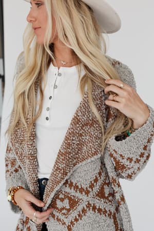 A woman with long blonde hair wearing a white hat, a white shirt, and the Free to Roam Cardigan in gray, featuring an Aztec-inspired print, poses holding her hair and showing off her rings and bracelets.
