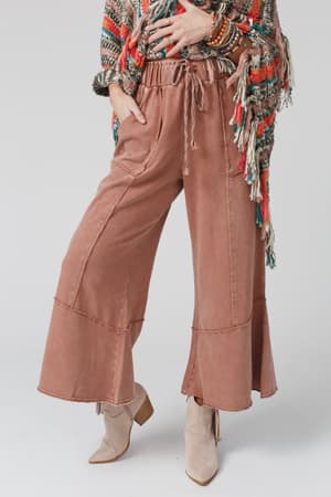 A person wears the Feeling Good Wide Leg Pant in Faded Rust with a drawstring waist, paired with a colorful fringed sweater, stacked bracelets, and beige heeled ankle boots, posing against a plain background.