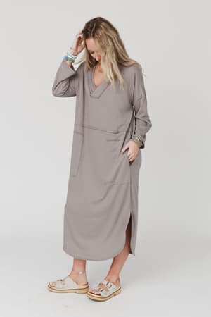 A woman wearing The Nest Meadow Long Sleeve Slit Maxi Dress - Coco, featuring large front pockets, stands against a plain background. She looks down, touching her hair, and finishes her look with sandals and layered bracelets.