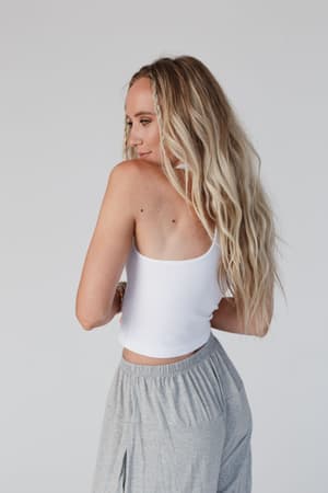 A woman with long, wavy blonde hair models the white High Neck Racerback Brami, paired with gray pants, as she stands sideways and looks over her shoulder against a plain light background.