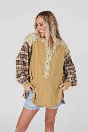 A woman smiles in the Better Together Mixed Print Top - Mustard, a loose long-sleeve featuring yellow floral and brown plaid designs, paired with light denim shorts against a plain white background.