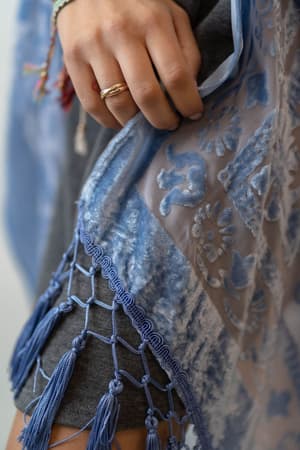 A close-up of a hand holding the edge of the Crushin' On You Velvet Kimono in Light Blue, featuring a patterned, semi-sheer design with tassels. The person wears rings, colorful bracelets, and a grey garment underneath.