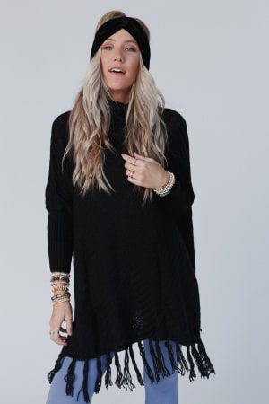 A woman with long, wavy blonde hair wears the Paloma Poncho Sweater in black with a headband, blue pants, and bracelets, standing against a plain light background.