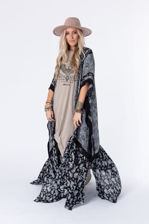 A woman with long blonde hair stands against a white background, wearing the Paisley Tapestry Free Flow Duster Kimono in Black over a graphic T-shirt dress, accessorized with a wide-brimmed hat and multiple bracelets.