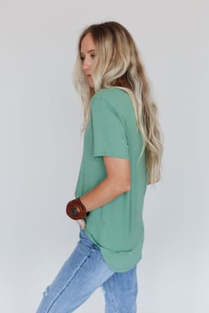 A woman with long, wavy blonde hair stands sideways against a plain white background, wearing the Scoop Neck Bralette Tee in Sage with light blue jeans and a brown bracelet.