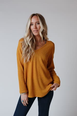 A woman with long blonde hair wears The Nest Flattering Open Back Thermal Knit Top in mustard and dark jeans, standing against a plain light background. She looks at the camera with a relaxed expression, holding the sweater’s hem.