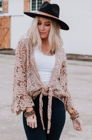 A woman with long blonde hair, styled in boho fashion, wears a black wide-brim hat, white top, Crochet Petal Kimono Wrap - Mocha tied at the waist, black pants, and bracelets while standing outdoors before a white building.