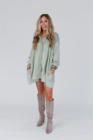 A woman with long blonde hair smiles against a plain background, wearing the Sunday Kind Of Love Dress in sage, styled with multiple bracelets and knee-high taupe suede boots.
