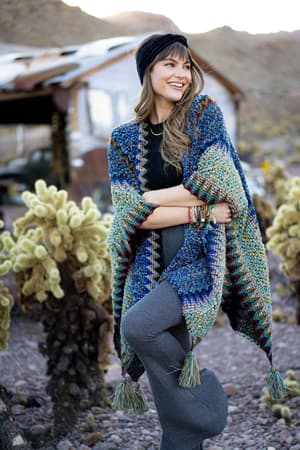 A woman stands outdoors near cacti and a rustic building, smiling with arms crossed as she wears the Desert Dreams Crochet Ruana - Cobalt, styled with a black headband and gray pants—a perfect addition to any boho closet.