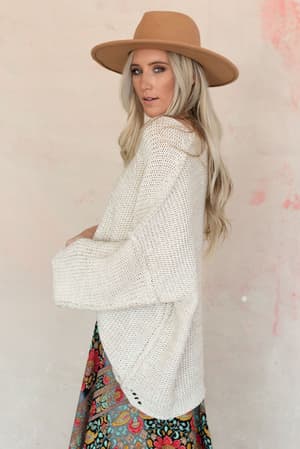 A woman with long blonde hair, wearing a tan wide-brimmed hat, the Crisp Air Sweater in Oatmeal, and a colorful floral skirt stands against a light pink and beige textured wall.