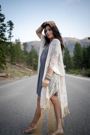 A woman stands confidently on a forested mountain road, posing in the Crushin' On You Velvet Kimono - Nude over a gray dress. The clear sky and tall trees create a peaceful, natural backdrop for her stylish look.