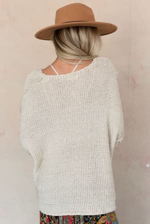 A woman with blonde hair in a low bun wears the Crisp Air Sweater in oatmeal, paired with a tan wide-brim hat and a colorful skirt. She stands with her back to the camera against a light beige wall.